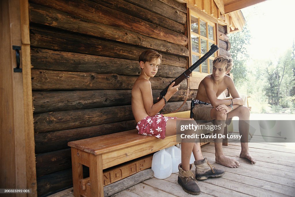 Teenage boy (13-15) and boy (10-13) sitting on bench in porch, boy holding rifle