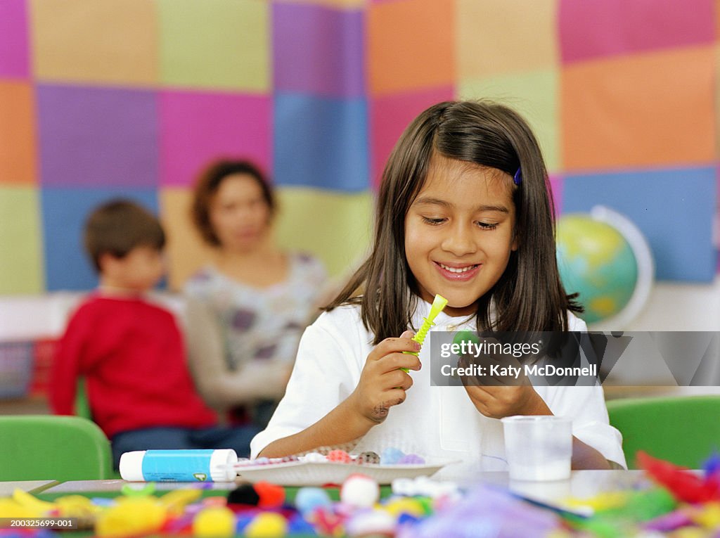 Schoolgirl (5-7) at table, applying glue to craft material, smiling
