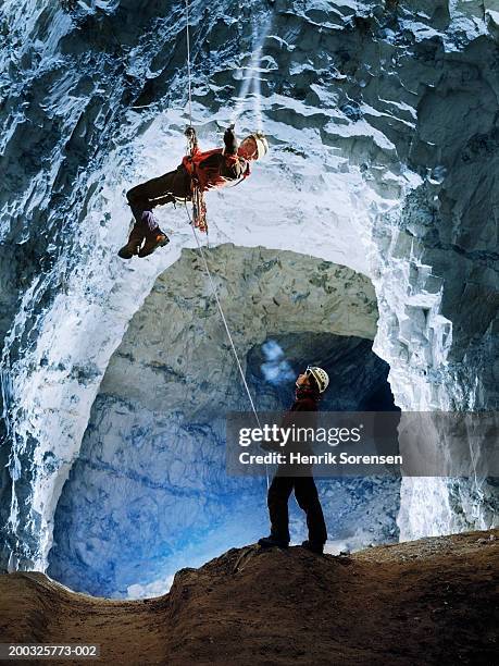 woman standing beneath mature man abseiling in cave, holding torch - espeleología fotografías e imágenes de stock