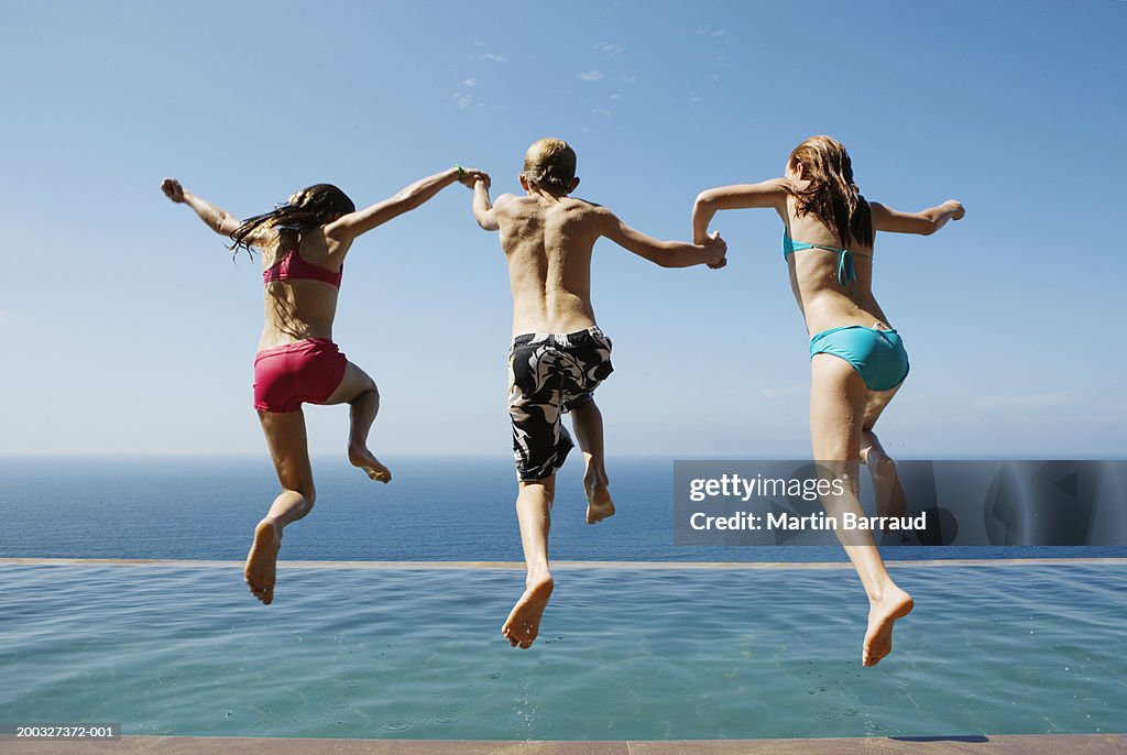 Two girls and boy (10-13) holding hands, jumping into pool, rear view