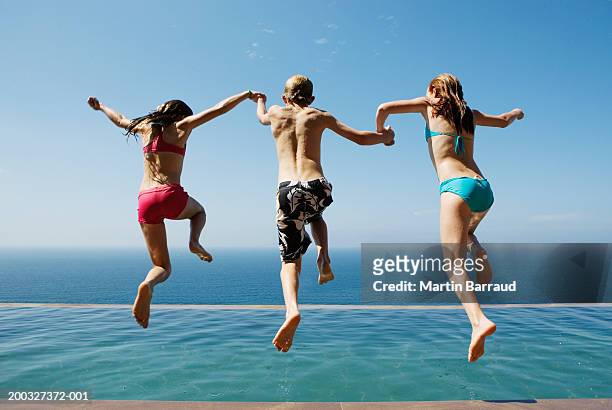 two girls and boy (10-13) holding hands, jumping into pool, rear view - jump in pool stock pictures, royalty-free photos & images