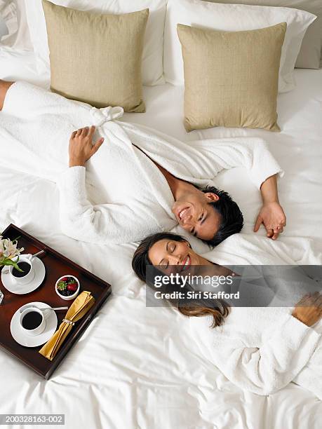 couple in white bathrobes lying on bed, breakfast tray on side - robe stock pictures, royalty-free photos & images