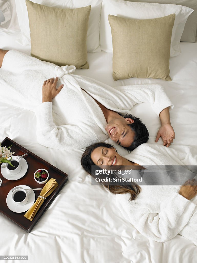 Couple in white bathrobes lying on bed, breakfast tray on side