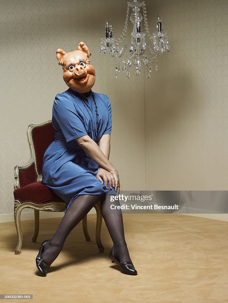 Businesswoman wearing pig mask sitting under chandelier, portrait
