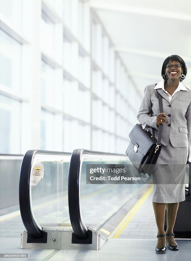 Woman pulling suitcase on moving walkway in airport, smiling