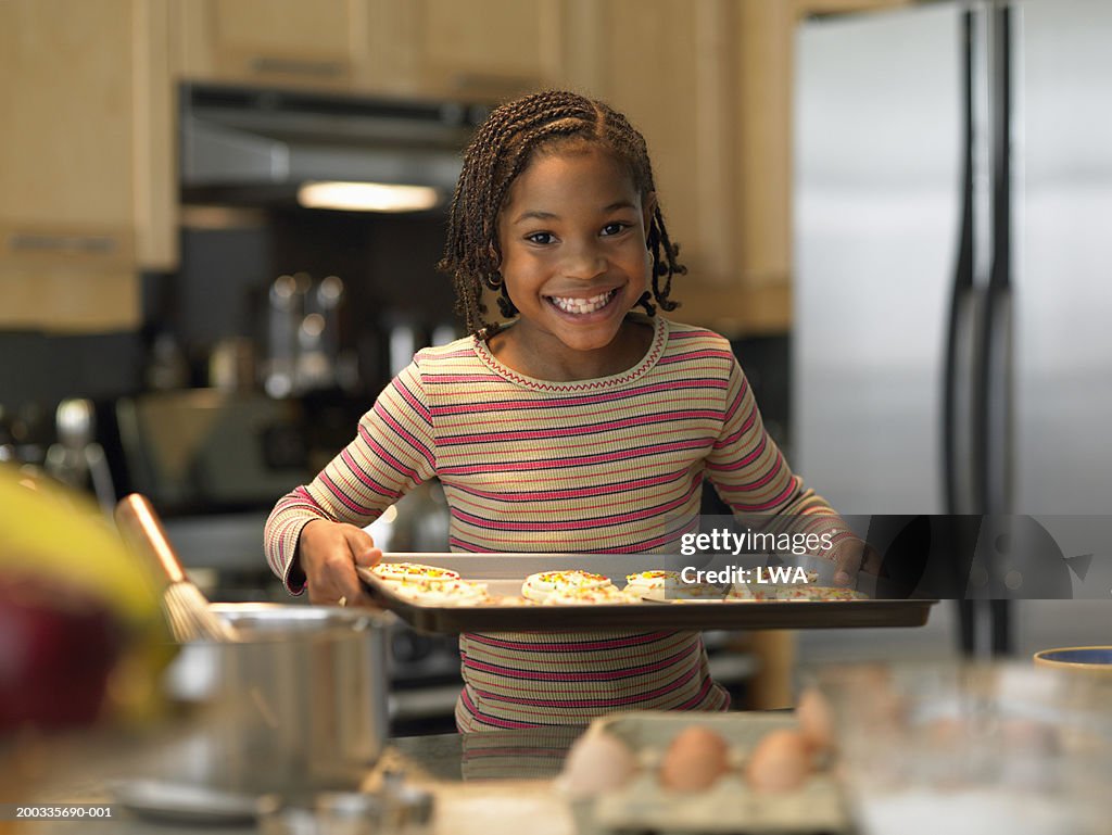 Girl (6-8) with tray of cookies in kitchen, smiling, portrait