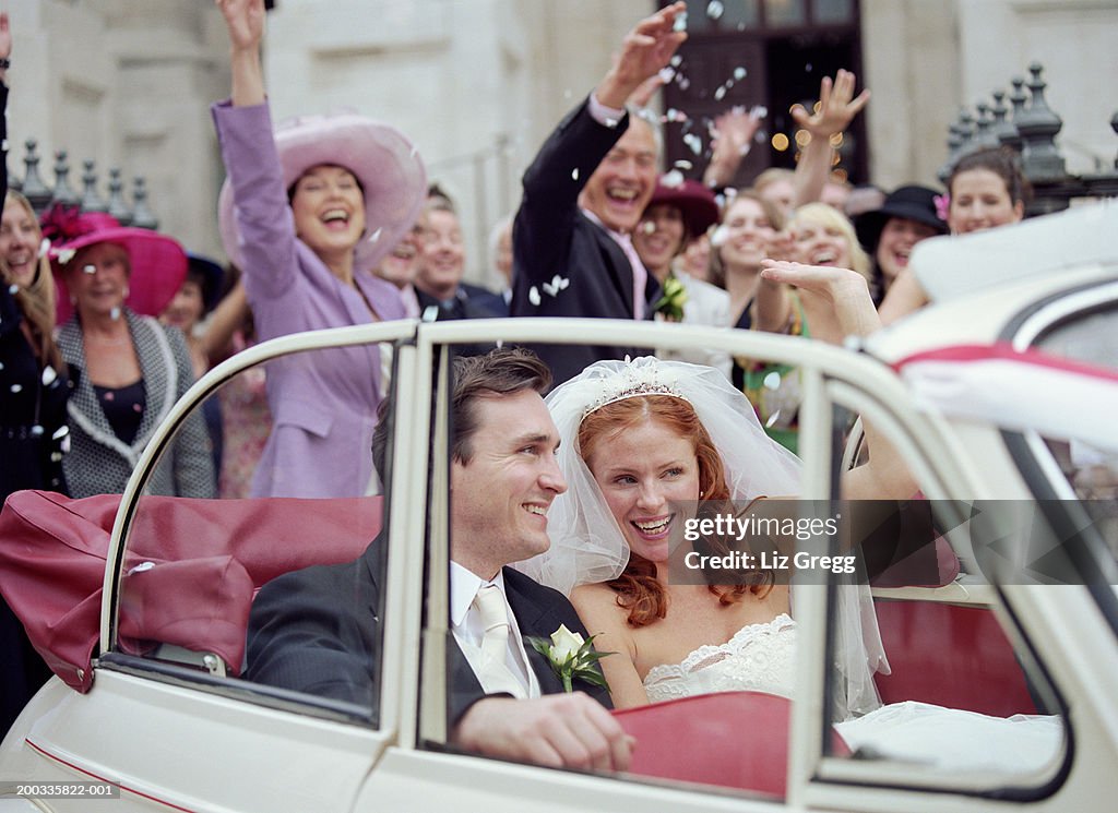 Bride and groom in convertible car, wedding party waving in background