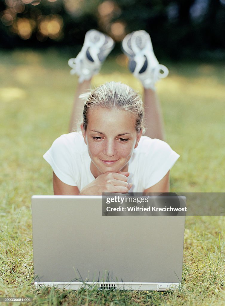 Young woman lying on grass by laptop, smiling