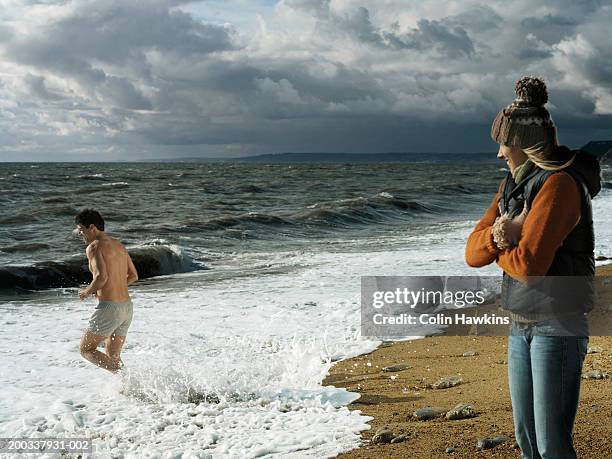 woman in woolly hat on beach watching man running into sea - couple winter stock pictures, royalty-free photos & images