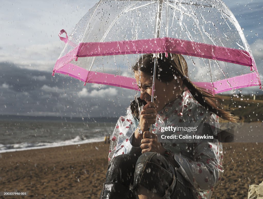 Girl (7-9) on beach, sheltering from rain under umbrella
