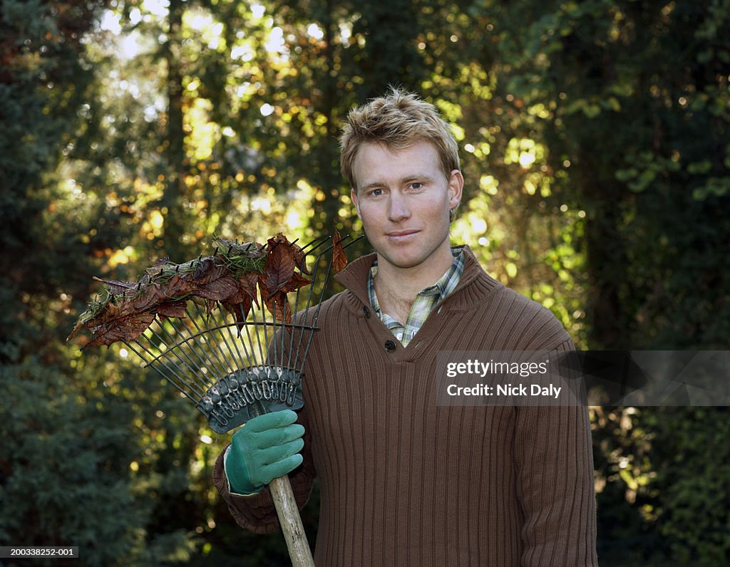 Young man standing in garden holding rake, smiling, portrait