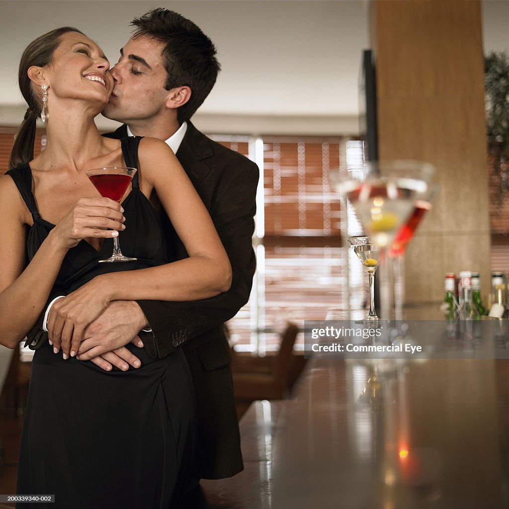 Man kissing woman's cheek from behind, woman holding cocktail