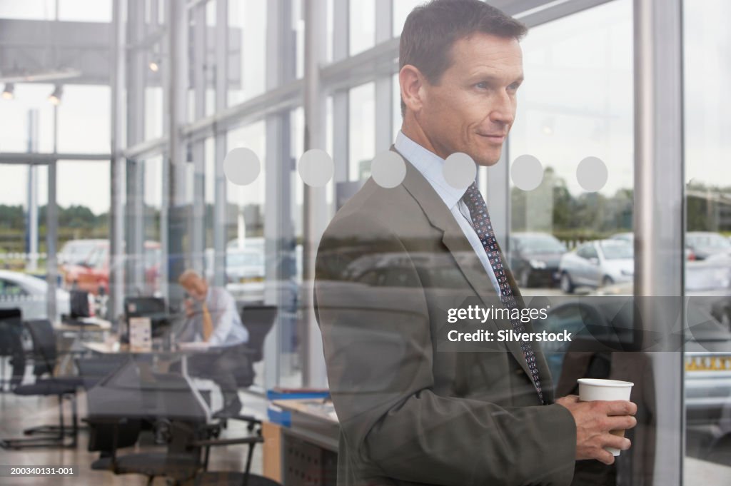 Businessman in car showroom holding drink, smiling, view through glass