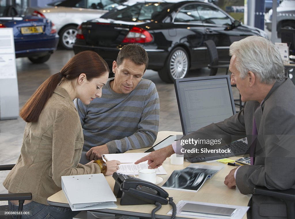 Couple sitting at desk signing document, mature car salesman pointing