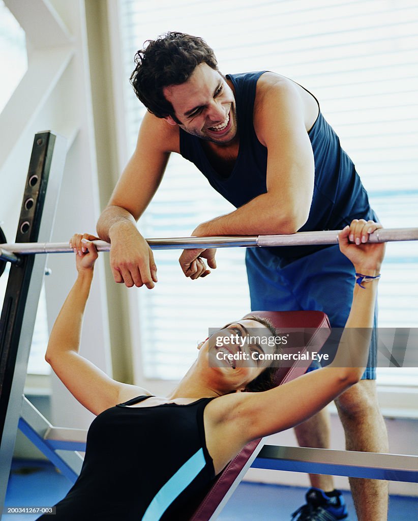 Woman sitting on weight bench, holding barbell, man leaning behind