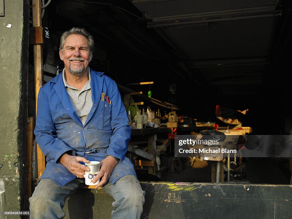 Senior male boat builder in workshop holding mug, smiling, portriat