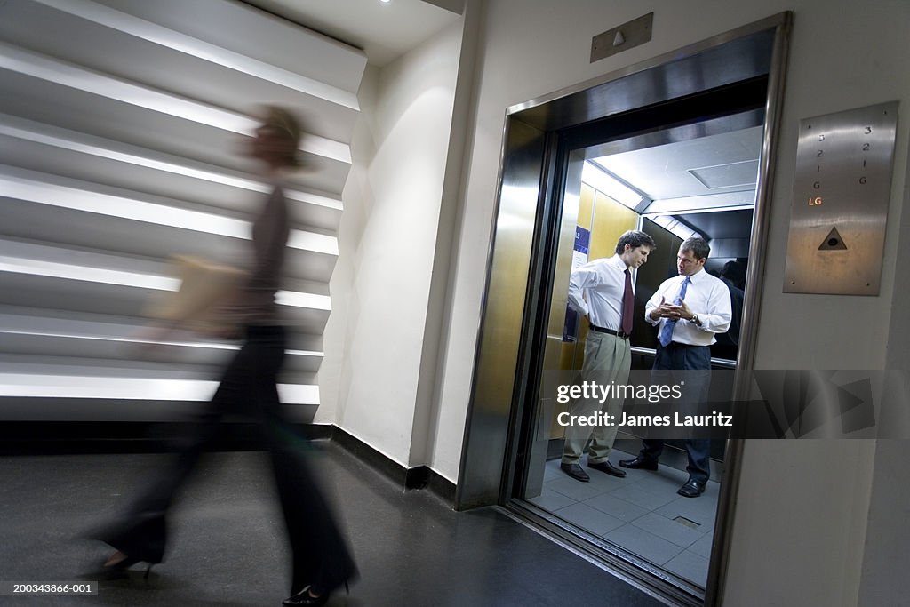 Two businessmen talking in elevator, woman passing (blurred motion)