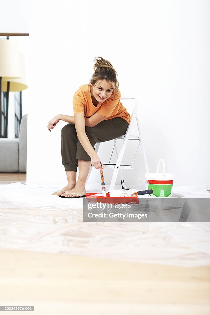 Woman sitting on step ladder, holding paint brush, smiling, portrait