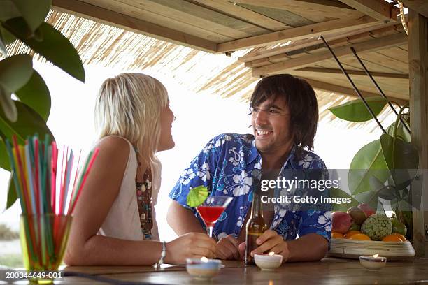 young couple sitting at beach bar, smiling at each other - bar de plage photos et images de collection