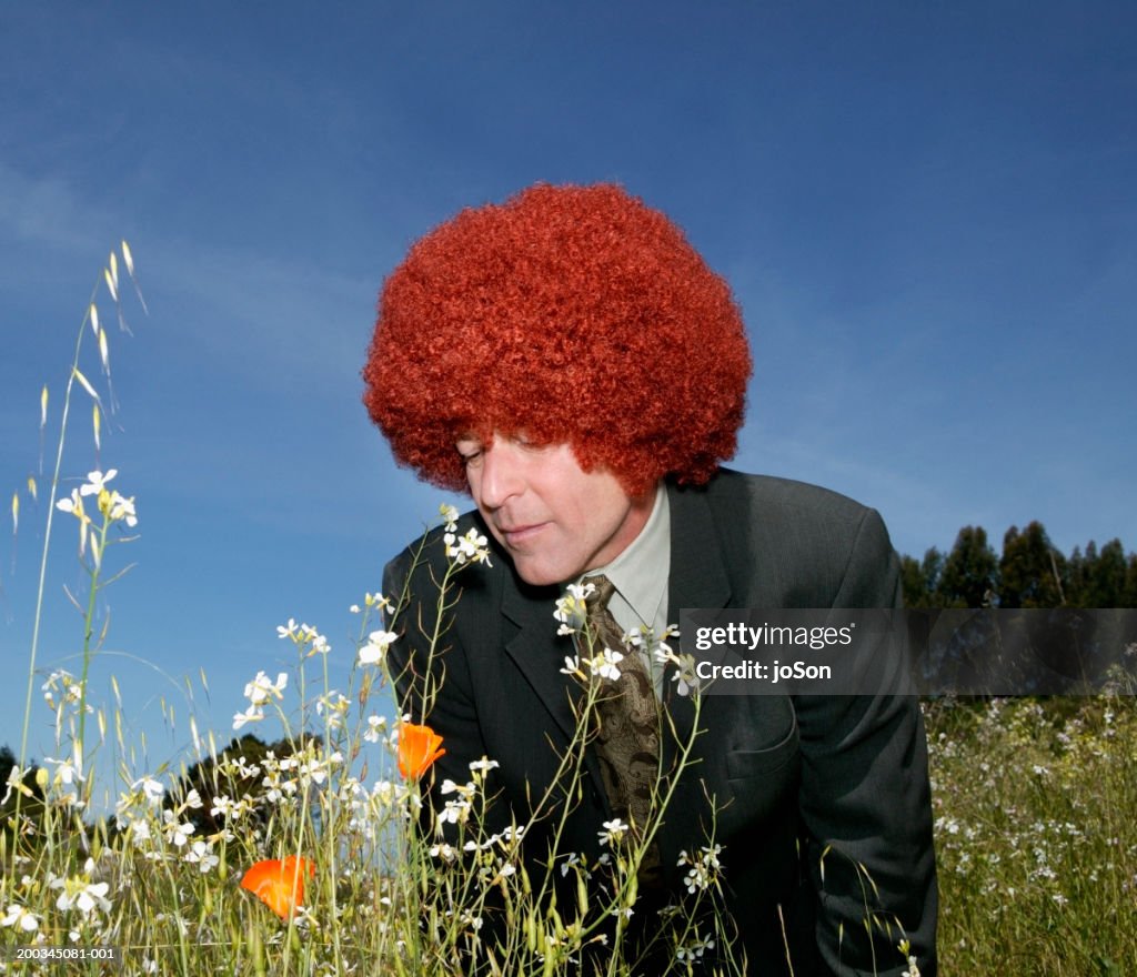 Mature businessman in red afro wig smelling flowers