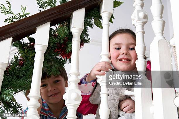 girl and boy (5-7) on stairs, looking through bannisters, smiling - christmas tree stockfoto's en -beelden