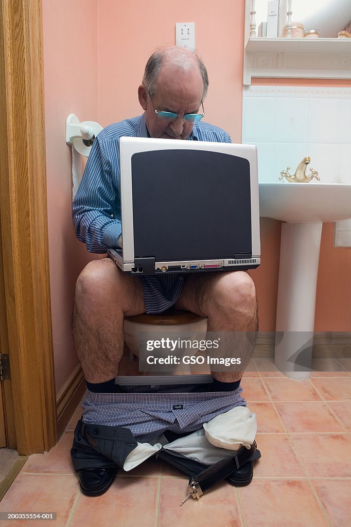 Senior businessman sitting on toilet, using laptop