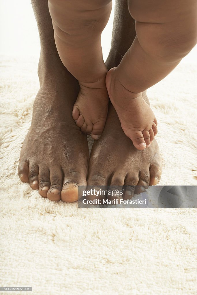 Baby girl (6-9 months) standing on man's feet, low section