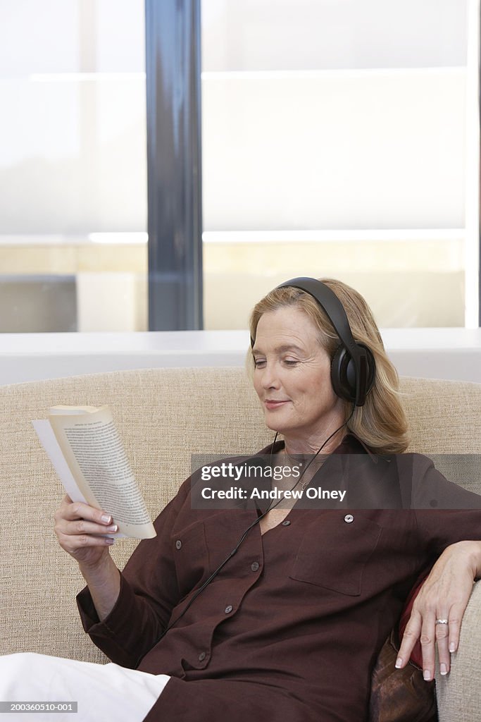 Mature woman sitting on sofa wearing headphones while reading book