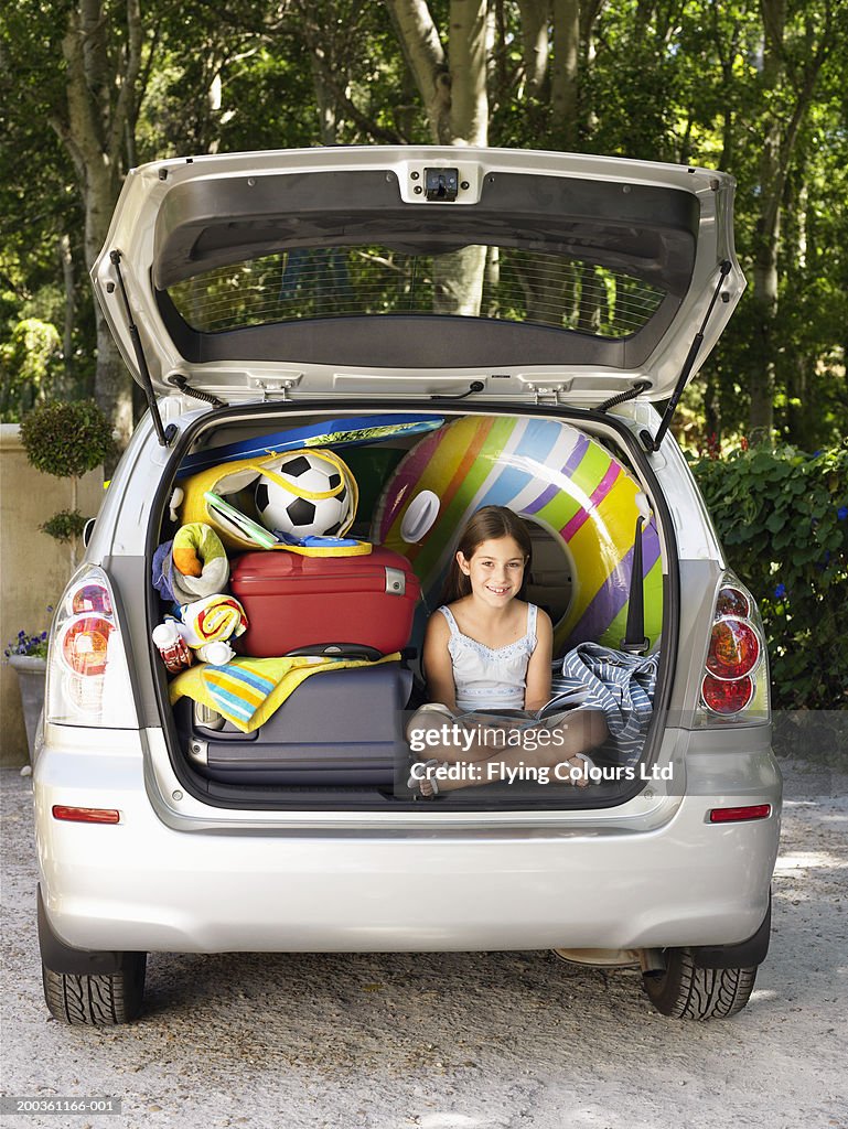 Girl (8-10) sitting in boot of car reading amongst luggage, portrait