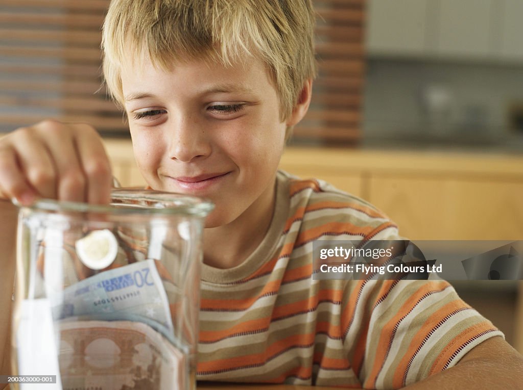 Boy (7-9) holding coin in money jar, smiling, close-up