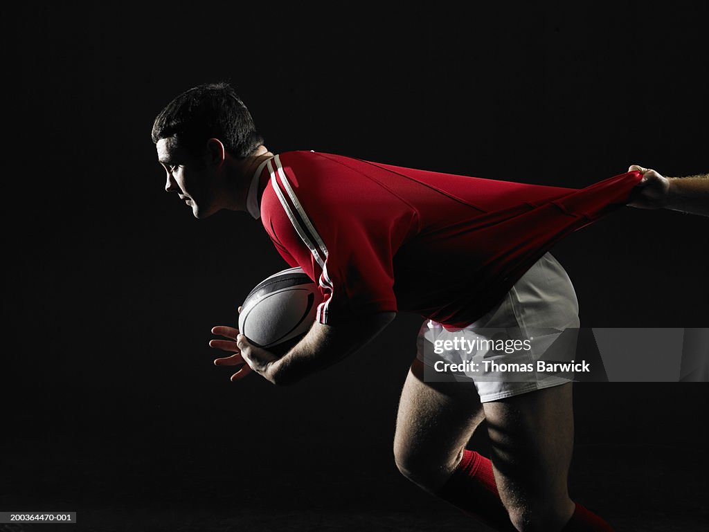 Two young men playing rugby, one carrying ball, one pulling on jersey