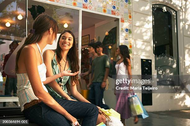 two young women sitting outdoors by shop, smiling - girl after shopping stock-fotos und bilder