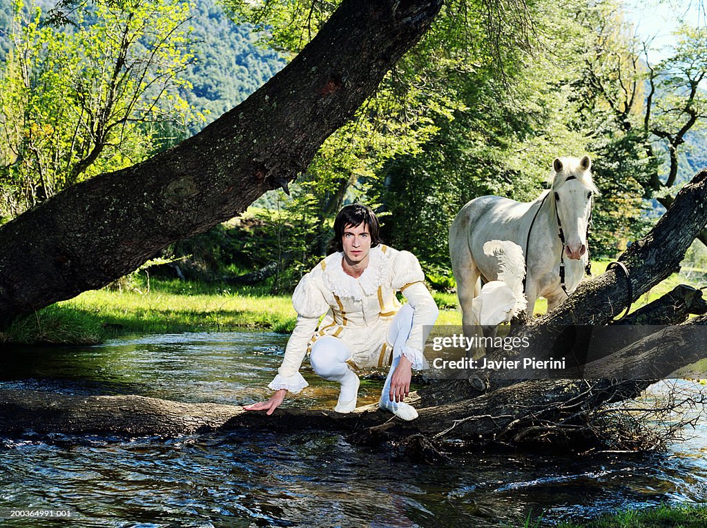 Young man in prince costume crouching by river, horse in background
