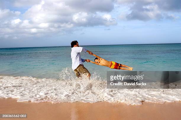father swinging son (5-7) in ocean, side view - swimming shorts stock pictures, royalty-free photos & images