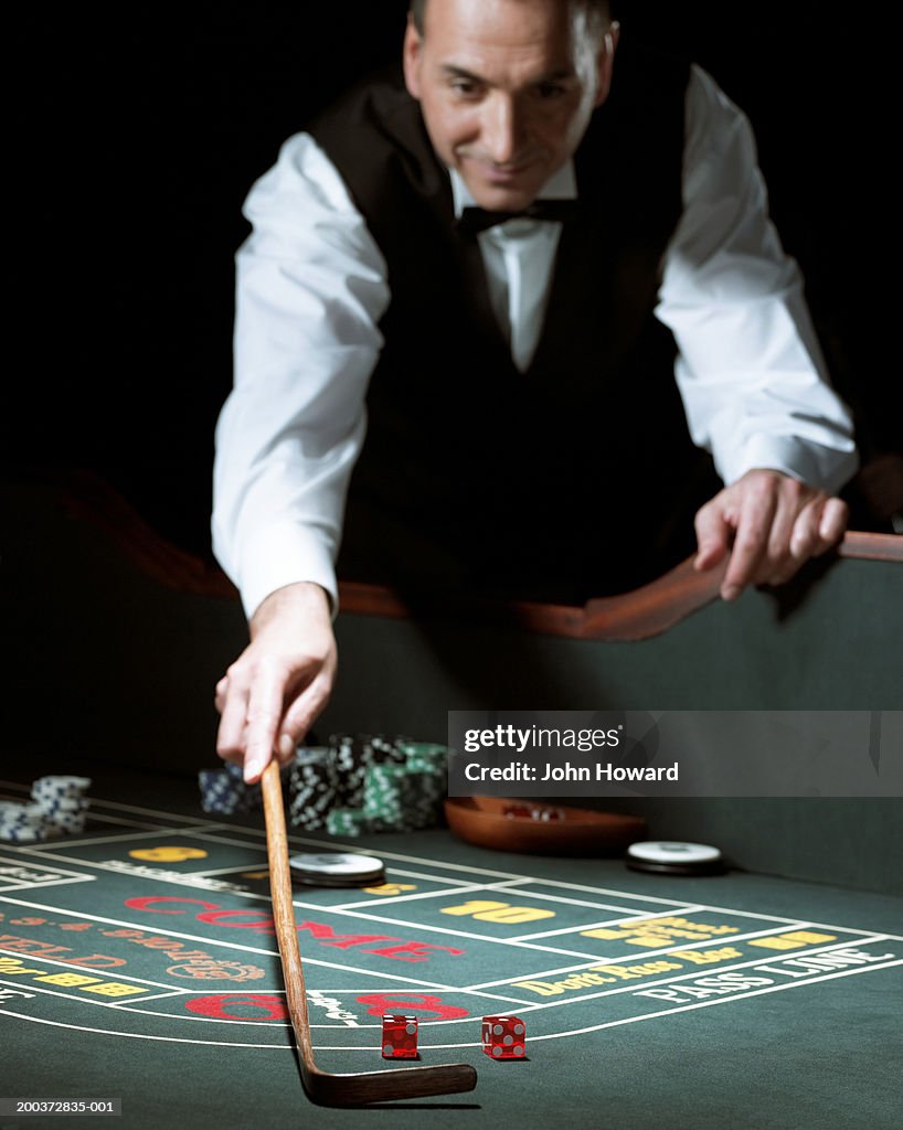 Male croupier leaning on gaming table using rake to retrieve dice