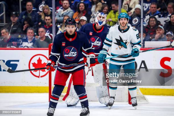 Goaltender Connor Hellebuyck of the Winnipeg Jets looks around a screen set by teammate Neal Pionk and Mike Hoffman of the San Jose Sharks during...