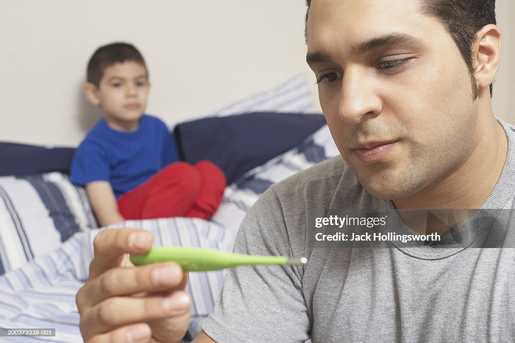 Close-up of a young man holding a digital thermometer with his son sitting on the bed behind him