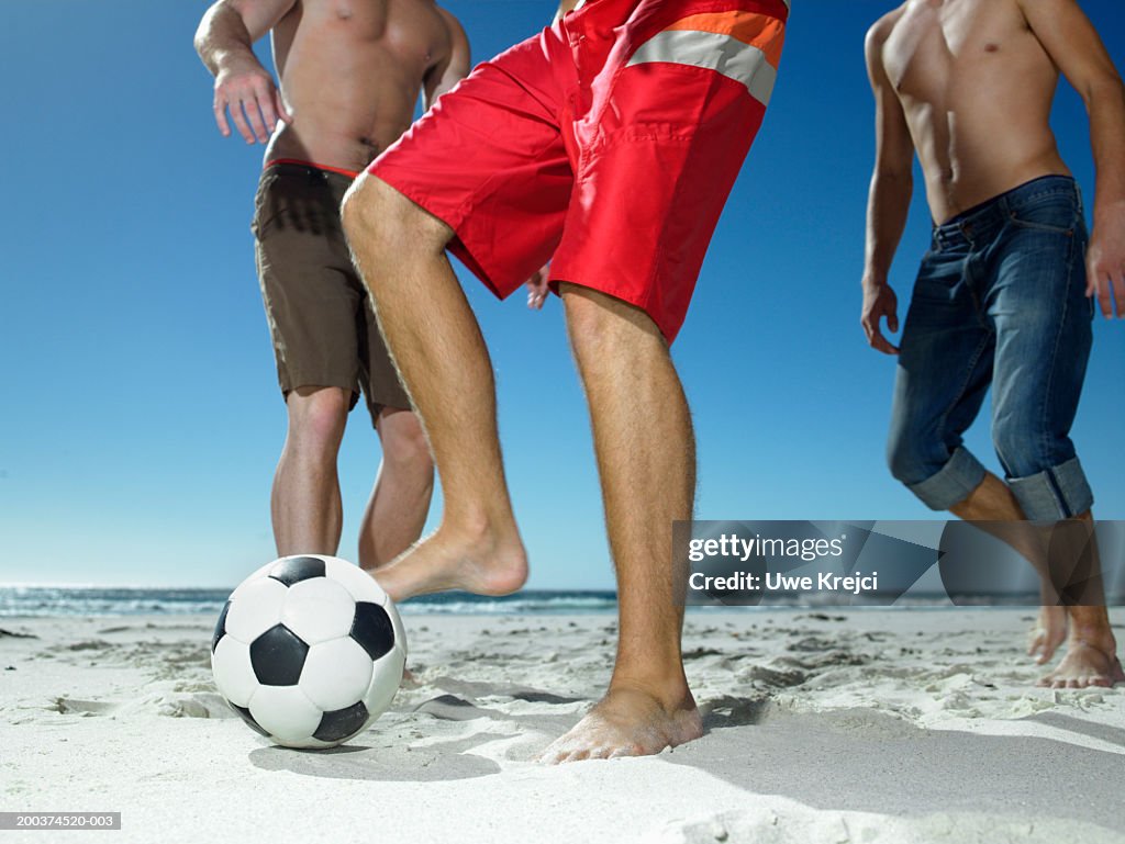 Three young men playing football on beach, low section