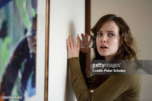 young woman using glass to listen through wall - entrometido fotografías e imágenes de stock