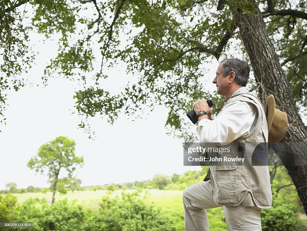 Senior man standing by tree holding binoculars, side view