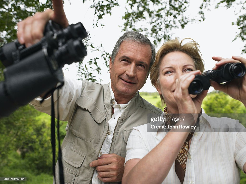 Mature couple holding binoculars, man pointing ahead, smiling