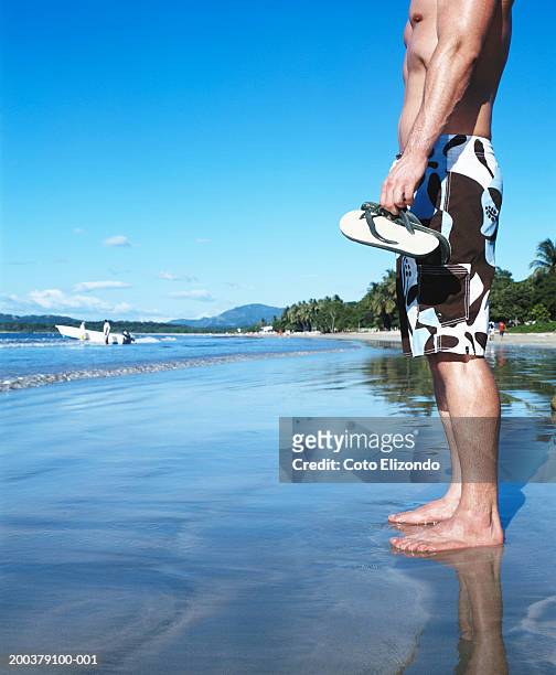 man in swim trunks standing on beach, low section, side view - playa tamarindo - fotografias e filmes do acervo