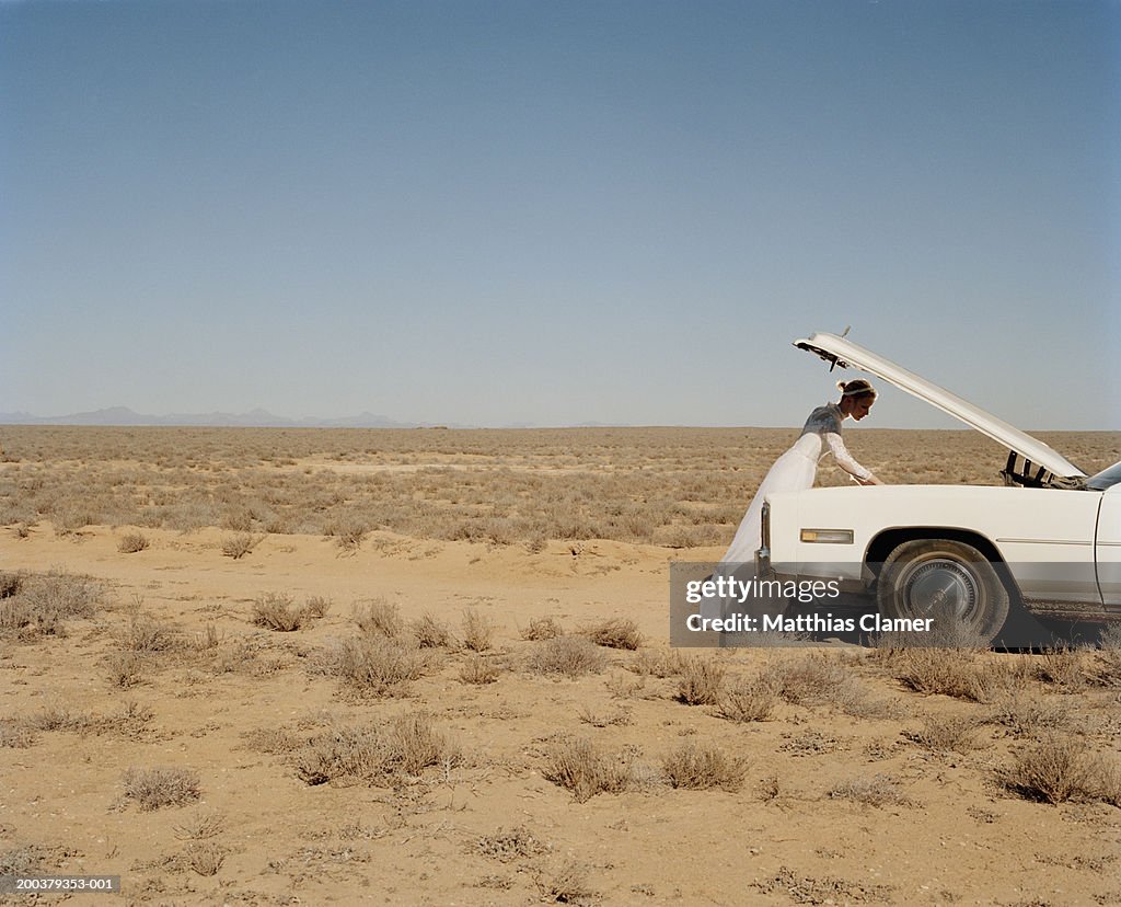 Bride looking under hood of stalled car in desert, side view