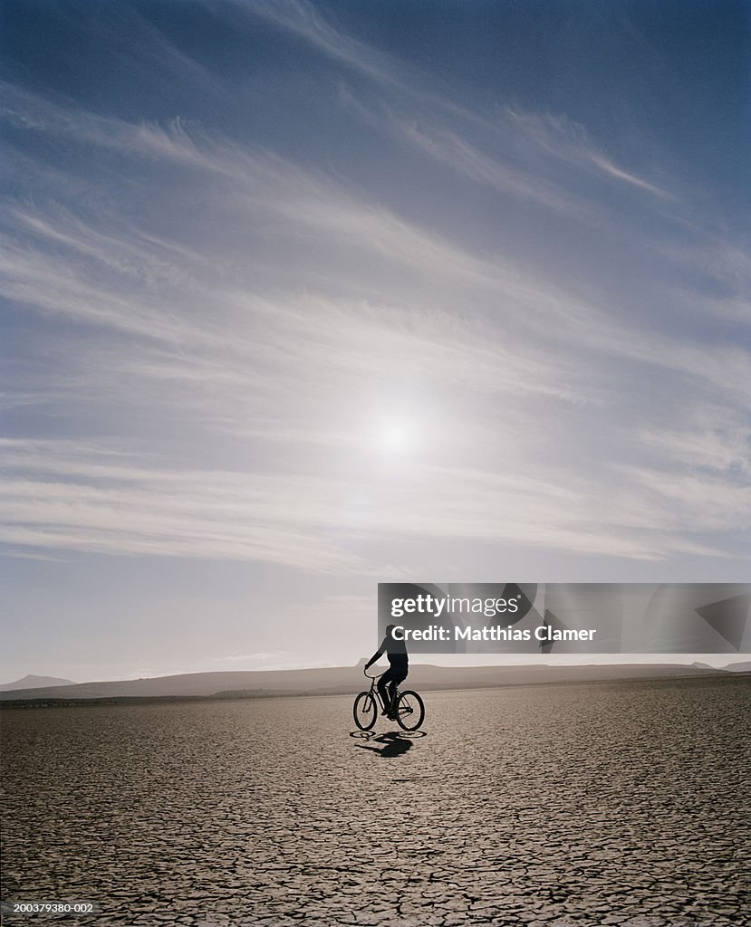 Sun shining above young woman riding bicycle in desert, side view