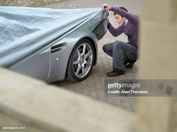 young man looking under protective sheet on car, smiling - covered car photos et images de collection