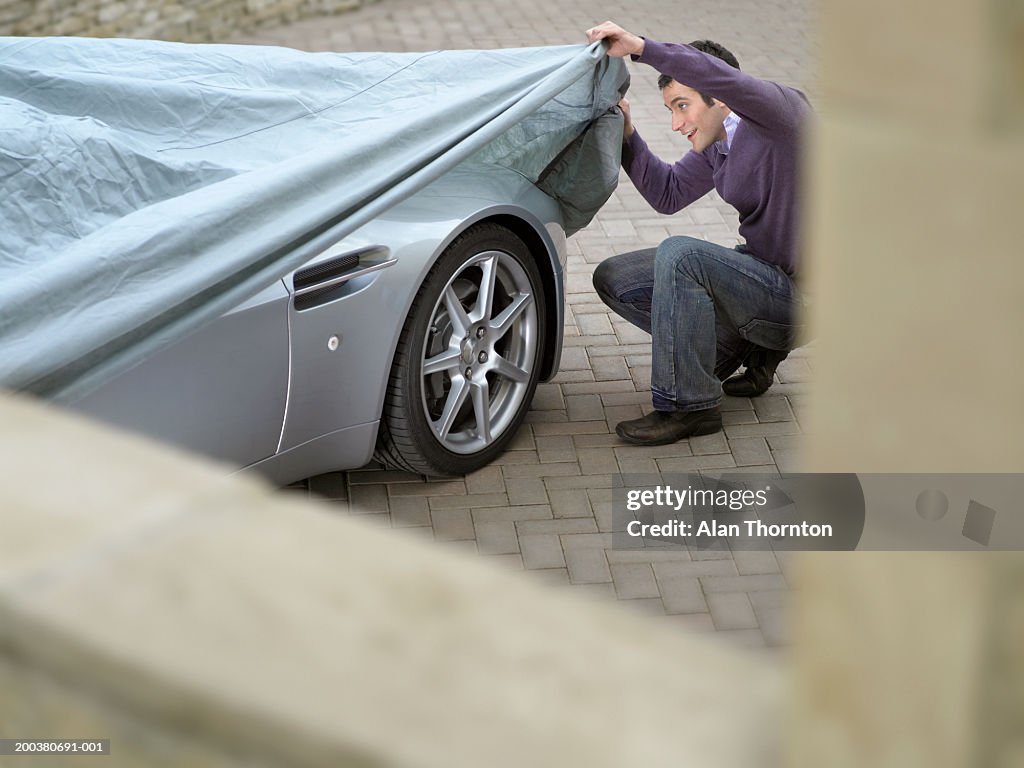 Young man looking under protective sheet on car, smiling