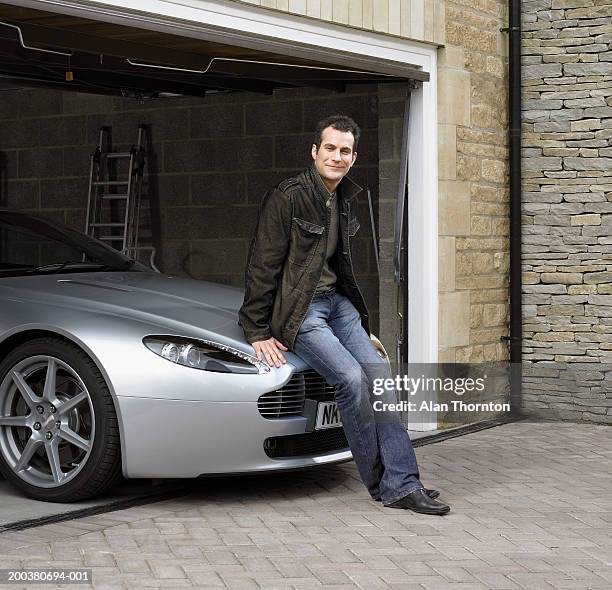 young man sitting on car in garage, smiling, portrait - silberner mantel stock-fotos und bilder