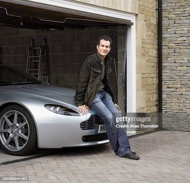 young man sitting on car in garage, smiling, portrait - luta fysisk ställning bildbanksfoton och bilder