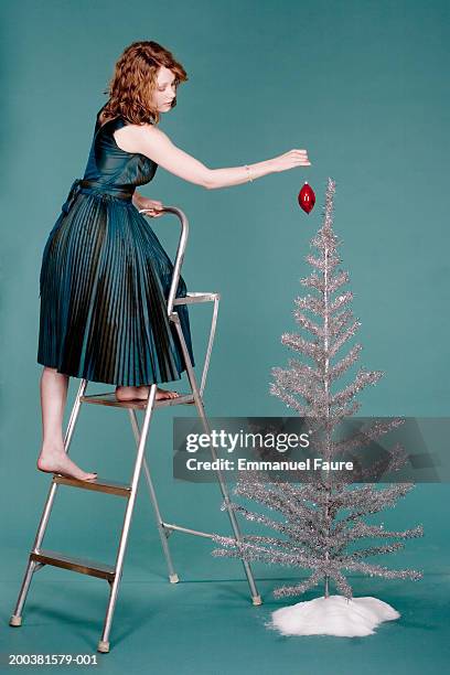 young woman on ladder putting christmas decoration on artificial tree - christmas tree stockfoto's en -beelden
