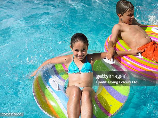 brother and sister (9-12) sitting on inflatables in swimming pool - rubber ring - fotografias e filmes do acervo